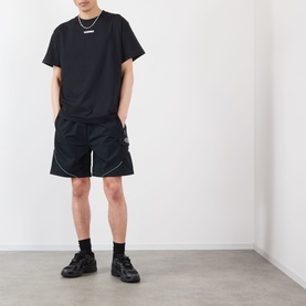 Model wearing Pleasures black cotton T-shirt with black shorts, white contrast-stitch detail, and black chunky trainers