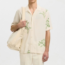 Model wearing Selected Casual Shirt in Egret with green floral print, short sleeves, and camp collar, styled with matching trousers and tote bag.