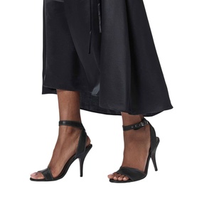 Model wearing All Saints black heeled sandals with ankle strap and stiletto heel, styled with black wide-leg trousers.