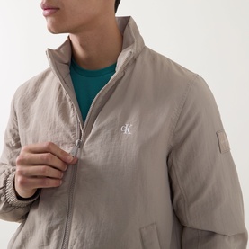 Model wearing Calvin Klein Jeans Harrington Logo Weatherproof Jacket in Vintage Khaki, with teal crew-neck tee visible beneath.