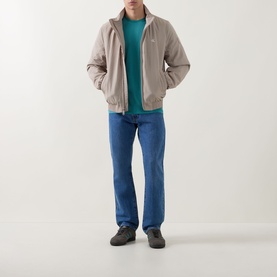 Model wearing Calvin Klein Jeans Harrington Logo Weatherproof Jacket in Vintage Khaki, styled with teal crewneck, blue jeans and grey trainers.