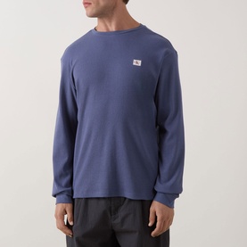 Model wearing Calvin Klein Jeans Waffle Knit Logo T-Shirt in Blue Nomad, long sleeves, crew neck, chest logo badge, styled with dark trousers.