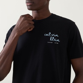 Model wearing Calvin Klein Jeans Travel Line Logo T-Shirt in black, crew neck with light blue script logo on chest.