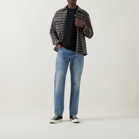 Model wearing Calvin Klein Jeans Travel Line Logo T-Shirt in black, styled with grey plaid overshirt, light blue jeans and black trainers.