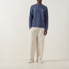 Model wearing Calvin Klein Jeans Badge Logo Sweatshirt in Blue Nomad, crew neck, long sleeves, styled with cream wide-leg trousers and white trainers.