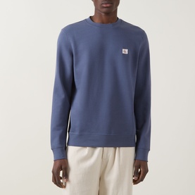Model wearing Calvin Klein Jeans Badge Logo Sweatshirt in Blue Nomad, crew neck with badge logo, paired with cream trousers
