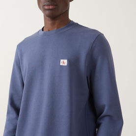 Model wearing Calvin Klein Jeans Badge Logo Sweatshirt in Blue Nomad, crew neck with small woven badge logo on chest.