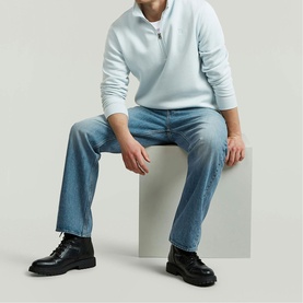 Model wearing G-Star baby blue half-zip sweatshirt, styled with light wash jeans and black chunky lace-up boots, seated on white cube.