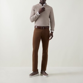 Model wearing Meyer brown cotton trousers, styled with a beige V-neck jumper, white shirt, brown belt and grey trainers.
