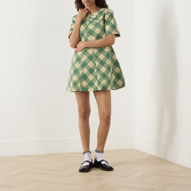 Model wearing Damson Madder Bridget Reversible Mini Dress in green gingham, A-line silhouette, short sleeves, styled with black Mary Janes and white