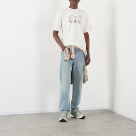 Model wearing Pellador Dog Tail Knot Logo T-Shirt in white with light wash jeans and grey trainers, jacket tied around waist.