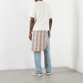Model wearing Pellador Dog Tail Knot Logo T-Shirt in white, styled with blue wide-leg jeans and a plaid overskirt, green trainers visible.