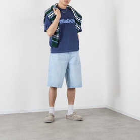 Model wearing Pellador Roundstone Logo T-Shirt in navy, styled with light wash denim shorts, white socks and grey trainers. Green striped jacket over