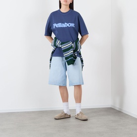 Model wearing Pellador Roundstone Logo T-Shirt in navy, styled with light blue denim shorts, striped jacket tied at waist, and beige sandals.