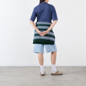 Model wearing Pellador Roundstone Logo T-Shirt in navy, seen from behind, styled with striped knit wrap, light blue shorts and beige sandals.