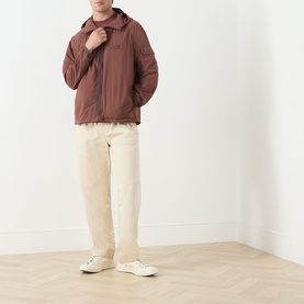 Model wearing Barbour Re-Engineered Flyweight Showerproof Jacket in Pepper brown with hood, styled with cream trousers and white trainers.