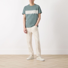 Model wearing Barbour Brayworth Graphic Logo T-Shirt in Vintage Teal with cream chest stripe, styled with off-white trousers and white trainers.