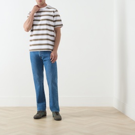 Model wearing Barbour white crew neck T-shirt with brown horizontal stripes, styled with blue straight-leg jeans and grey trainers.