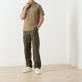 Model wearing Barbour Milburn Graphic Logo T-Shirt in Clay, styled with olive cargo trousers and white trainers.