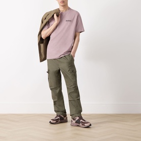 Model wearing Barbour Moonwell Graphic Logo T-Shirt in Ironstone pink, styled with olive cargo trousers, tan jacket and mauve trainers.