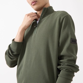Model wearing Barbour International Half Zip sweat in Thyme green, textured cotton fabric with quarter zip collar and logo badge sleeve.