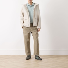 Model wearing Barbour polo in Dusty Slate, styled with a cream hooded jacket, taupe trousers and grey trainers.