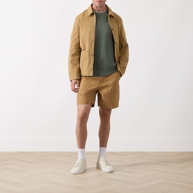 Model wearing Barbour Sherburn Logo Straight-Leg Shorts in Camel with a khaki tee, tan jacket, white socks and cream trainers.