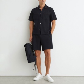 Model wearing Reiss blue short-sleeve shirt and matching shorts, carrying a navy backpack, styled with white trainers.