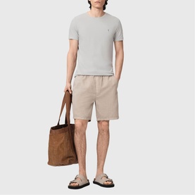Model wearing All Saints Tonic Ramskull Logo T-Shirt in Pebble Grey with beige shorts, styled with tan sandals and brown suede tote bag.
