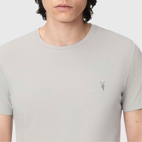 Model wearing All Saints Tonic Ramskull Logo T-Shirt in Pebble Grey, featuring crew neck and signature Ramskull logo embroidery at chest.