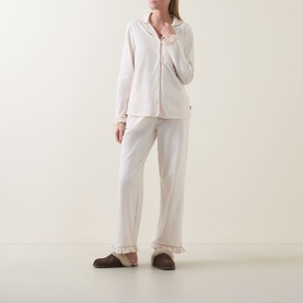 Model wearing Chelsea Peers Just Married Frill Long Pyjama Set in cream with frill cuffs, contrast piped trims, and brown slippers.