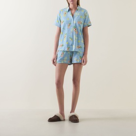 Model wearing Chelsea Peers Tennis & Cocktails Short Pyjama Set in blue print with collared top and shorts, styled with fluffy brown slippers