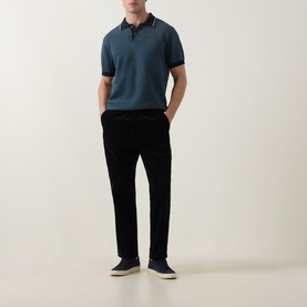 Model wearing Rodd & Gunn Oyster Island Striped Knitted Polo Shirt in teal, styled with black trousers and navy trainers.
