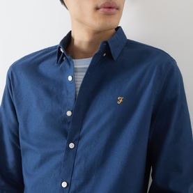 Model wearing Farah Vintage Brewer Logo Shirt in Aegean Blue, long sleeves, button-down collar, gold logo badge, styled with striped tee underneath.