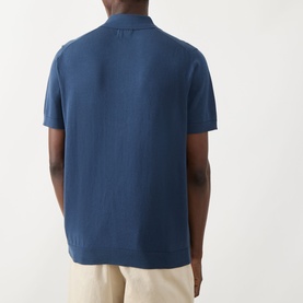Model wearing Farah Vintage Poplar Knitted Polo Shirt in Aegean Blue, short sleeves, polo collar, back view, styled with cream trousers.