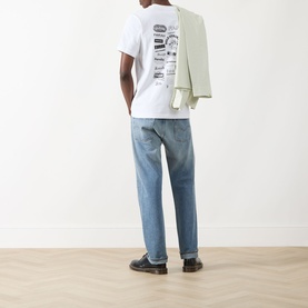 Model wearing Farah Vintage Logo Back Graphic T-Shirt in white with back graphic print, styled with light blue jeans and a sage blazer over shoulder.