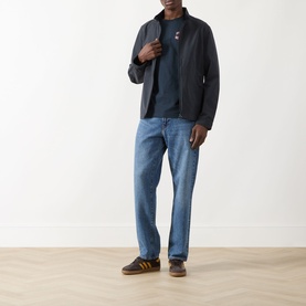 Model wearing Farah Vintage Caligo Logo T-Shirt in True Navy under dark zip jacket, styled with light blue jeans and dark trainers.