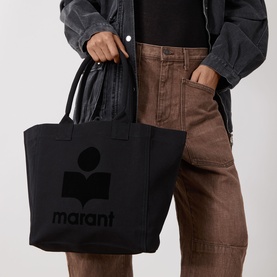 Model carrying Isabel Marant Yenky-GZ Small Tote Bag in black canvas with tonal logo print and top handles, styled with rust trousers