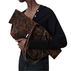 Model carrying All Saints bag in leopard brown, styled with a black top and dark jeans. Large clutch-style silhouette with suede-effect finish.