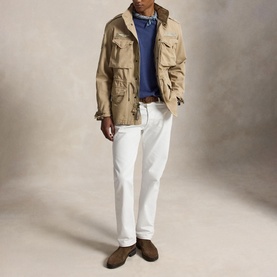 Model wearing a khaki field jacket with chest pockets, styled with a blue top, white trousers, and brown suede shoes