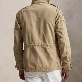 Model wearing khaki cotton field jacket, rear view, with epaulettes, waist drawstring, and pocket detail, styled with white trousers