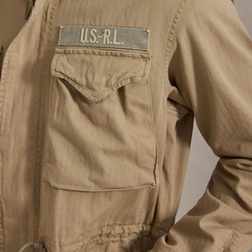 Close-up of khaki military-style jacket back, featuring U.S.R.L. embroidered patch on chest pocket, utility pocket below