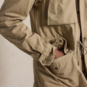 Model wearing a khaki utility parka with drawstring waist, chest pockets and snap-button cuffs, hand tucked into side pocket.