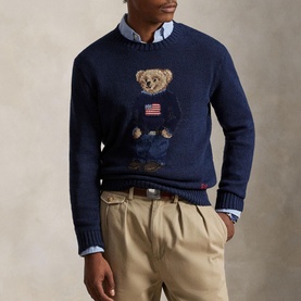 Model wearing navy knit crewneck sweater with Polo Bear holding American flag, styled with blue shirt and khaki trousers