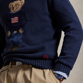 Model wearing navy Ralph Lauren knit sweater with embroidered Polo Bear and American flag motif, styled with khaki trousers and chambray shirt