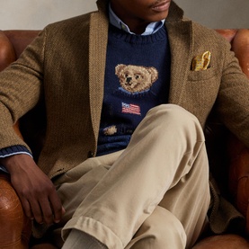 Model wearing navy Polo Bear knit sweater with American flag detail, layered under tan herringbone blazer with chino trousers