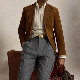 Model wearing brown corduroy blazer, cream cable-knit jumper, grey tailored trousers and gold belt, holding a brown leather bag.