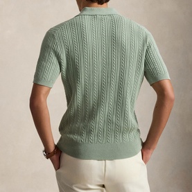 Model wearing sage green short-sleeve cable-knit polo shirt, rear view, styled with white trousers