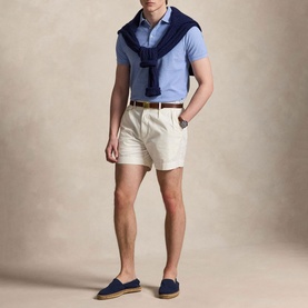 Model wearing light blue polo shirt and cream shorts, styled with navy sweater over shoulders, brown belt, and navy loafers