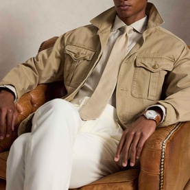 Model wearing Ralph Lauren Performance Twill Logo Shirt in Expedition Dune, styled with white trousers and a watch, seated in a leather chair.